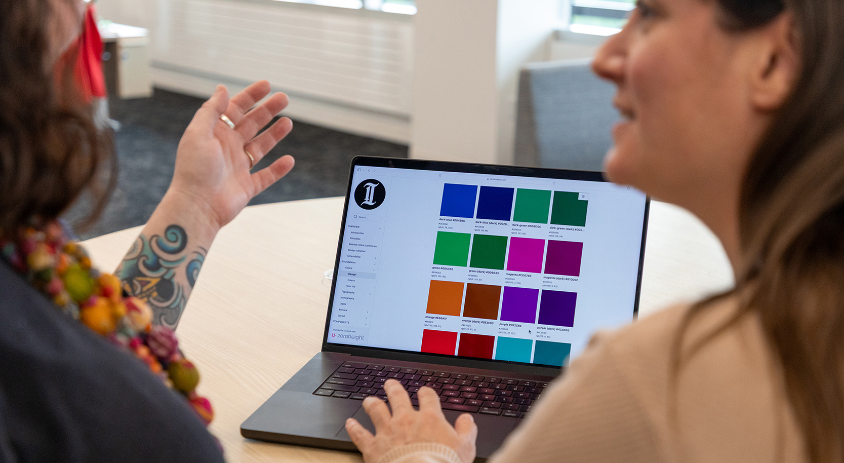 Two women sit at a round table reviewing a digital color palette labeled with hex codes on a laptop screen, which features branding from The Philadelphia Inquirer.